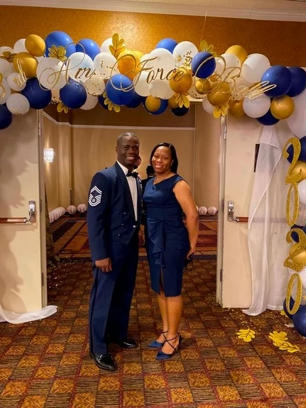 Chief Seals in service dress with his wife at a celebration under a blue and gold balloon arch.